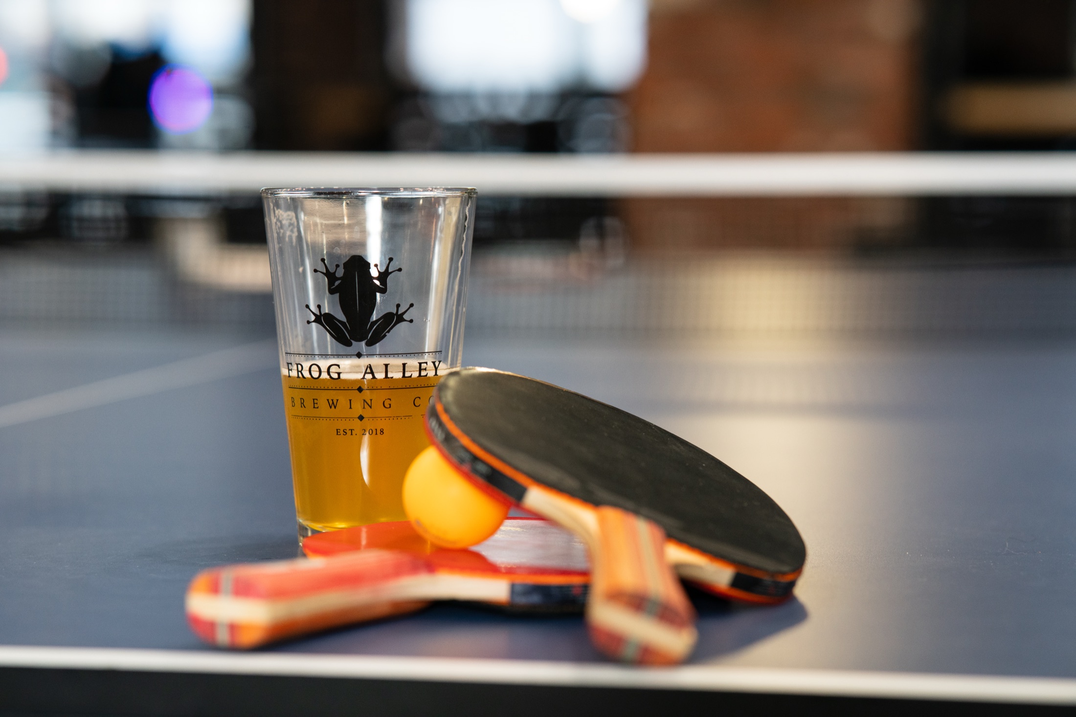 A Frog Alley Brewing pint glass and ping pong paddles on the table